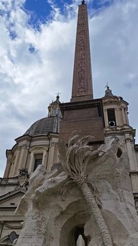 Fountan in Piazza Navona, featuring a lion sculpture and palm tree, with an ancient obelisk in the background, set against a historic architectural backdrop in Rome, Italy