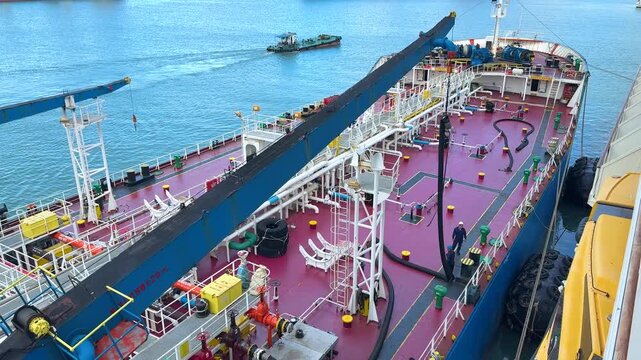 Workers are busy on the deck of a large tanker in a harbor. They operate machinery and handle equipment while the ship is docked. Tasks include loading and unloading cargo.