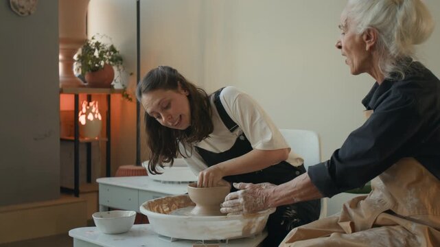 Young inexperienced woman failing shaping of clay bowl while elderly craftswoman correcting mistake at potters wheel in workshop