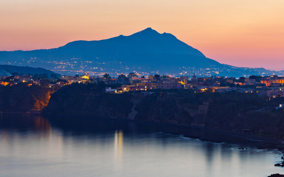 Long exposure evening photo of Procida with illuminated buildings on cliff above calm Mediterranean sea. Silhouette of Ischia island rises in background under sunset gradient sky