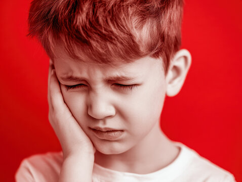 Portrait of a young boy with closed eyes holding his cheek experiencing toothache or pain against a vivid red background for health concept