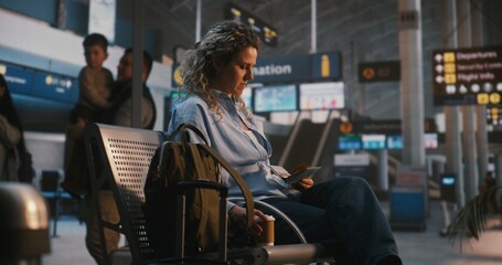 Obraz premium Female Traveler Arrives at Departure Hall With Backpack, Sits on Bench, and Compares Ticket With Information Board. Woman Takes Moment to Enjoy Coffee While Waiting For Boarding. Independent Travel.