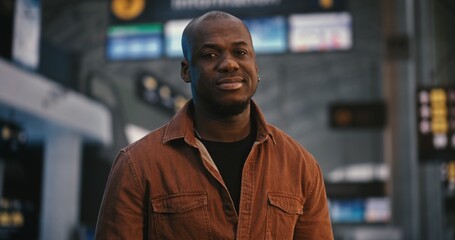 Fototapeta premium Portrait of African American Man Stands Peacefully in Center Crowded Departure Hall. While Other Passengers Move Quickly Around, Guy Remains Still and Composed, With Gentle Smile Playing on Lips.