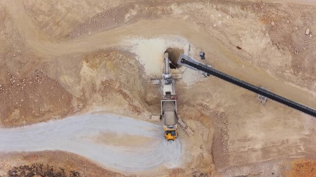 Aerial view of a quarry where heavy machinery loads earth onto trucks, contrasting the beige terrain with the dark conveyor, Arsoli, Lazio, Italy.