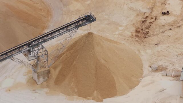 Aerial view of a sprawling sand pile under a metal conveyor belt, creating a textured landscape of light and dark tones, Arsoli, Lazio, Italy.