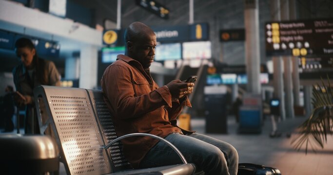 Black Male, Pulling Suitcase Enters Airport Waiting Area. Man Finds Seat and Uses Smartphone Scan Boarding Pass, Glances Between Device and Overhead Flight Information Boards Confirm at Correct Gate.