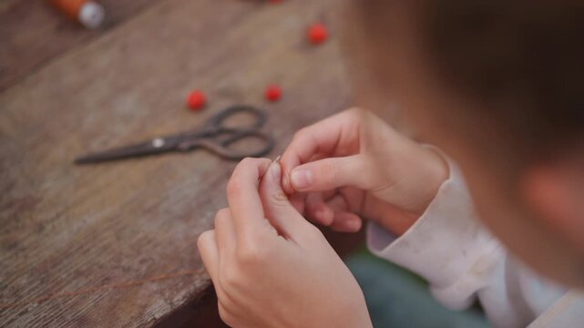 Closeup caucasian child threading beads. Playful outdoor craft moment on wooden bench with scattered red beads, scissors nearby, blurred face, nimble fingers assembling necklace, curiositydriven