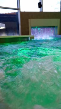 Green illuminated water bubbles and churns inside a modern indoor jacuzzi with a waterfall feature. Concept of hydrotherapy, relaxation, luxury wellness, and chromotherapy treatments.