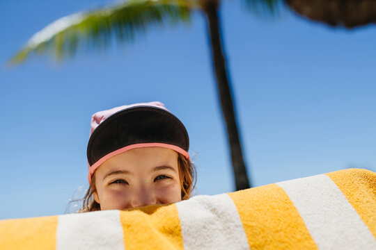 Smiling child peeking over beach towel under palm tree.