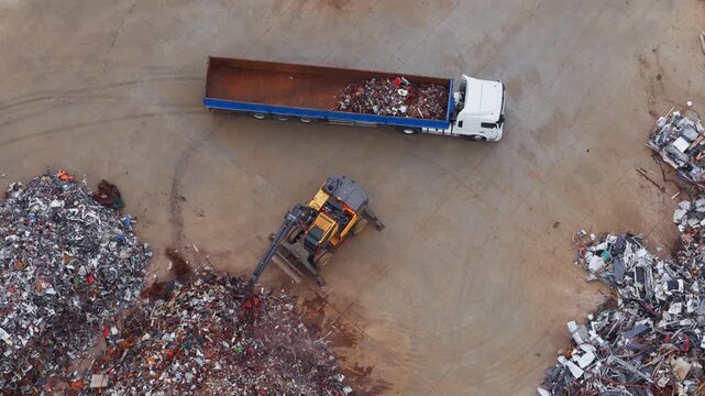 Aerial view of a yellow crane loading scrap metal into a truck, highlighting the stark contrast between the heavy machinery and the metallic debris, Avezzano, Abruzzo, Italy.