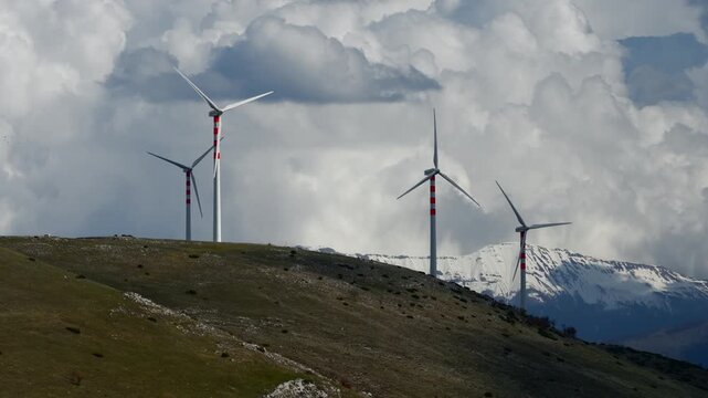 Aerial view of wind turbines standing tall against a backdrop of snow-capped mountains under a cloudy sky, Cocullo, Abruzzo, Italy.