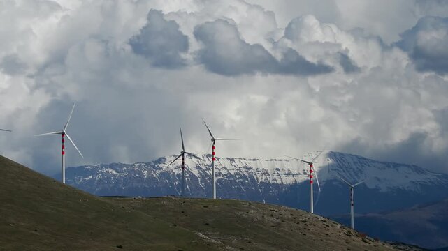 Aerial view of wind turbines standing tall on a green hill, with snow-capped mountains in the background, Cocullo, Abruzzo, Italy.