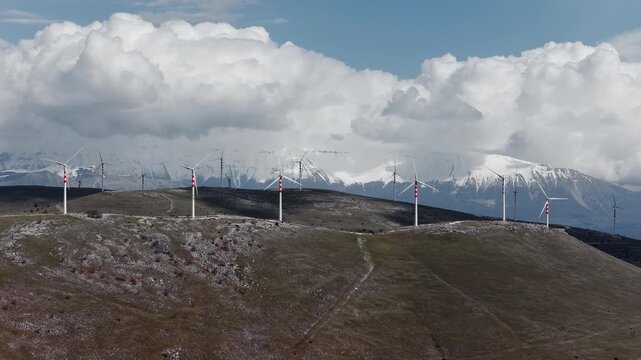 Aerial view of wind turbines standing tall on a hilltop against the backdrop of snow-capped mountains and a cloudy sky, Cocullo, Abruzzo, Italy.