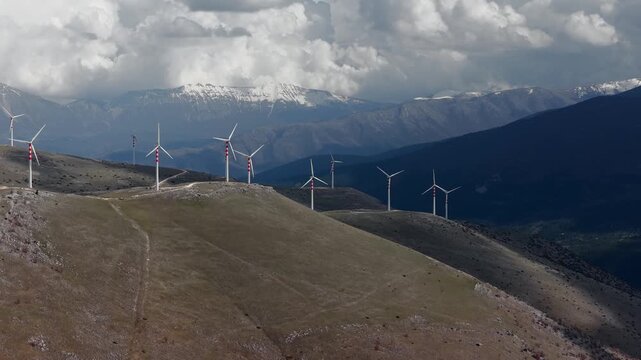 Aerial view of wind turbines standing tall on rolling hills, with snow-capped mountains in the distance under a cloudy sky, Cocullo, Abruzzo, Italy.