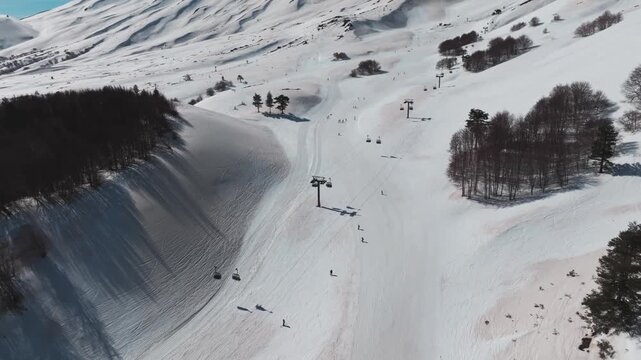 Aerial view of skiers descending the snowy slopes near dark forests, with chairlifts visible against the stark white landscape, Piano Provenzana, Sicilia, Italy.