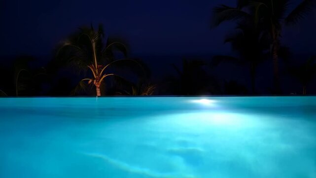 Static shot of illuminated infinity pool water gently rippling with palm trees silhouetted against night sky