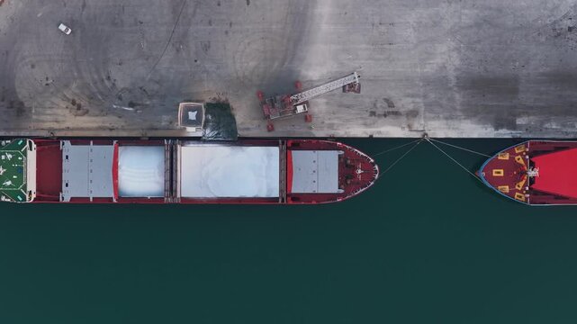 Aerial view of two cargo ships, one with white cargo, tied to a concrete pier, contrasting with the dark water, Vasto, Abruzzo, Italy.