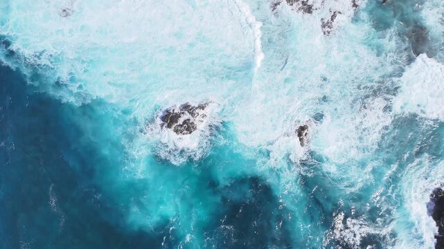 Turquoise ocean waves crash against rocky shoreline, creating foam and splashes, with vibrant blue water and white surf visible in coastal landscape. Aerial view to sea. Vibrant coastal scene