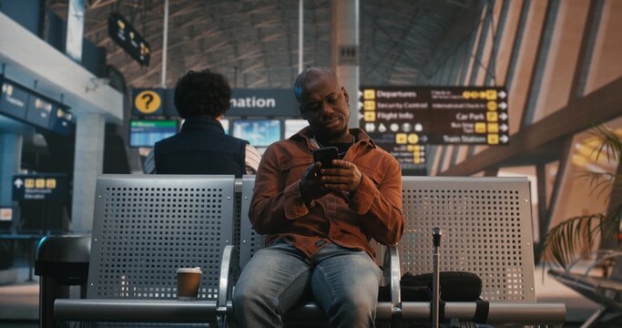 Young Black Man Arrives at Airport Waiting Area, Sets Down Luggage, Immediately Checks Smartphone. Modern Habit of Staying Connected, Checking Flight Updates, Chatting With Friends Before Departure.