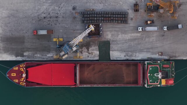 Aerial view of a large cargo ship docked at a port with cranes and trucks, contrasting the red interior with the blue-green water, Vasto, Abruzzo, Italy.
