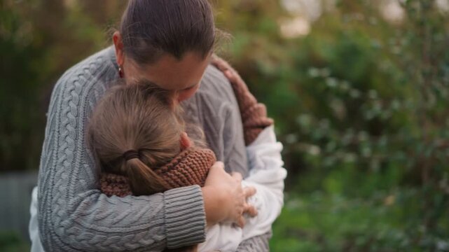 Caucasian mother hugging daughter outdoors, knit sweater and brown scarf, warm dusk light, garden foliage background, tender forehead kiss, protective arms, emotional reunion, serene atmosphere, close