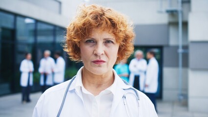 Fototapeta premium Charming Caucasian woman working as doctor. Relaxing outside hospital complex while smiling gently towards camera. Specialist holding relaxed posture. Medical staff in background discussing.