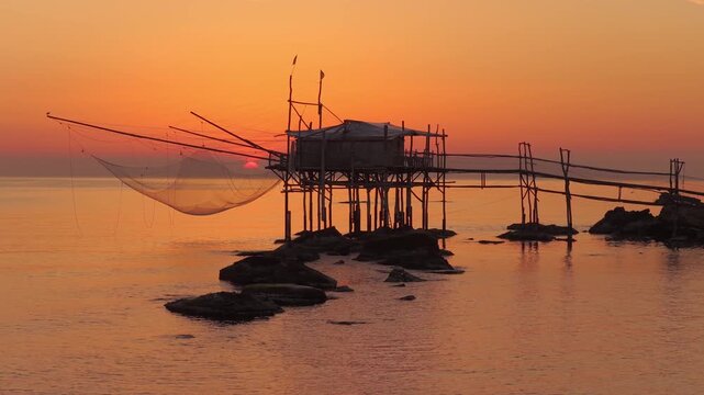 Aerial view of Trabucco, a traditional fishing structure, stands silhouetted against the fiery sunset hues over the Adriatic sea, San Vito Chietino, Italy.