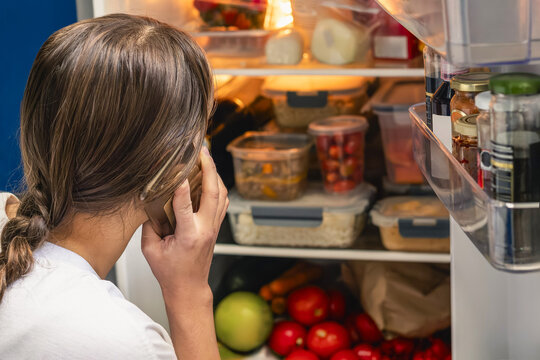Woman talking on mobile phone selecting food from refrigerator