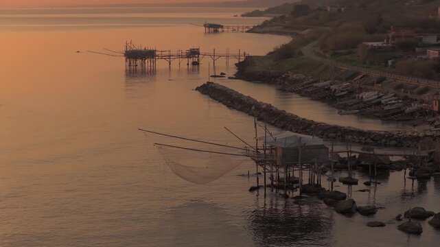 Aerial view of traditional Trabucchi fishing structures stand silhouetted against the sunset's golden glow creating a serene scene, San Vito Chietino, Abruzzo, Italy.