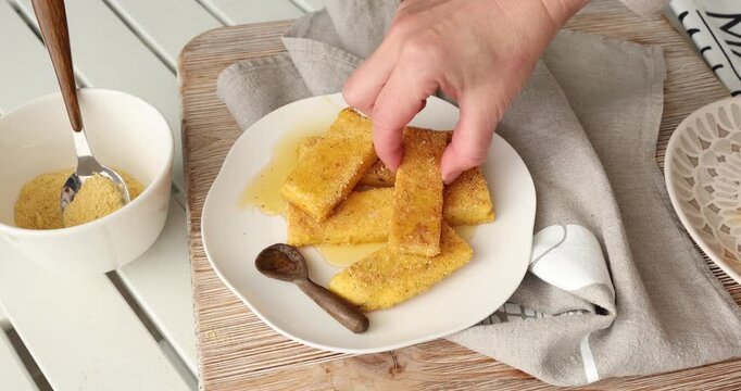 Hands pick up corn snack from plate. Crispy fried polenta sticks served on wooden board. Side dish in bright kitchen setting.