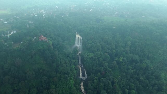 Aerial view of the Curugsewu Waterfall cascading through lush green forest with a building on a hill, Curugsewu, Central Java, Indonesia.