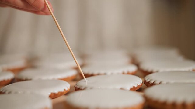 Closeup hand finishing iced cookies on tray using toothpick to create smooth glaze and tiny patterns, soft warm light, shallow depth of field, intimate kitchen bake studio, meticulous pastry work,