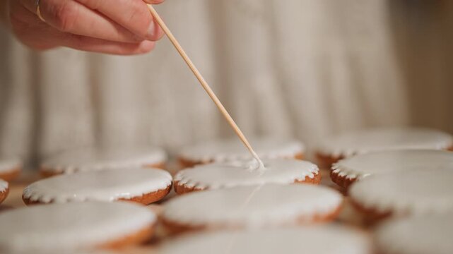 Macro caucasian hand precise pumpkin icing strokes over round cookies, focused pastry artisan creating uniform patterns with toothpick, studio kitchen light, meditative technique capturing texture