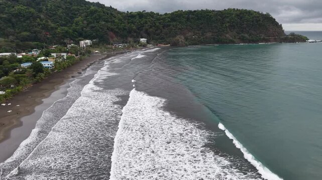 Aerial view of a dark sand beach where the waves crash against the shore near a lush green mountain, Jaco, Costa Rica.