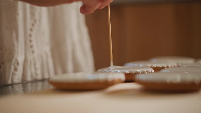 Closeup caucasian hand decorating pumpkin pie cookies on wooden table, slow homestyle baker using toothpick to shape white icing, warm soft light, cozy autumn kitchen mood, rustic tray and seasonal