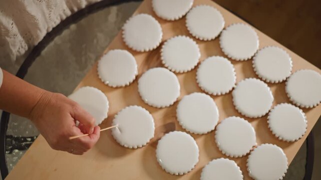 Caucasian hands decorating iced cookies on wooden board, closeup of precise toolwork smoothing fondant tops across rows of round treats, neutral warm light, artisanal baking routine and careful