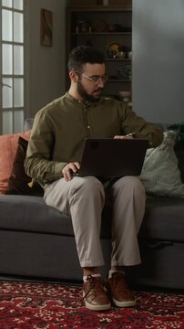 Vertical full length shot of adult Muslim man starting daily prayer at home checking watch while using computer then taking off shoes and laying out prayer rug on floor in modern living room