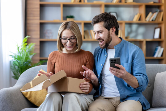 Cheerful couple on a modern sofa unboxing a home delivery, woman opening a parcel while man reacts with joy and smartphone in hand, enjoying the surprise and connection