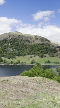 Elevated panoramic view footage of Rydal Water surrounded by green hills and trees under a blue sky with scattered clouds, Lake District, England, UK.