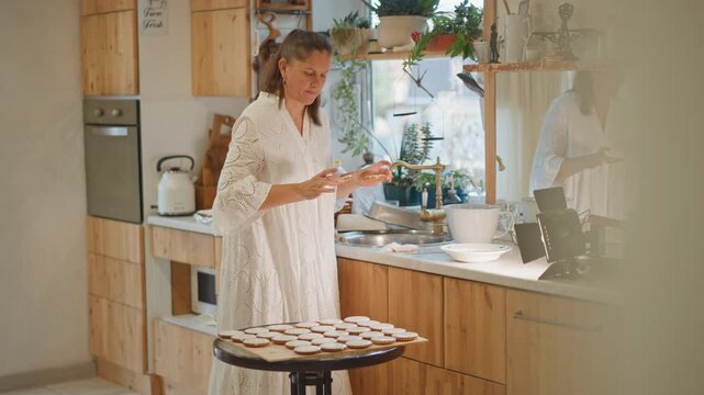 White woman arranging pumpkin pies, kitchen. Sunlit wooden counter, lace dress, cooling tray of pies, spice jars, flour dust, ceramic bowls and whisk, potted herbs by window, warm autumn tones,