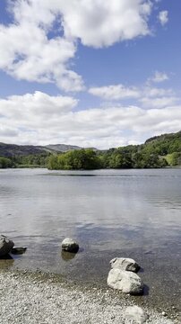 Shoreline footage of Rydal Water with scattered rocks, calm water and surrounding hills under a partly cloudy sky, Lake District, England, UK.