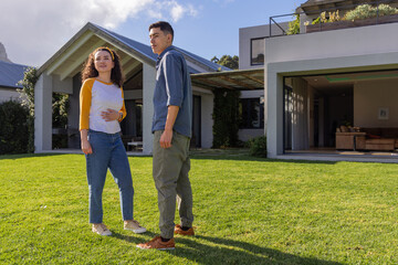 Fototapeta premium Couple standing on well-kept lawn in front of modern house, wearing raglan shirt and cargo pants