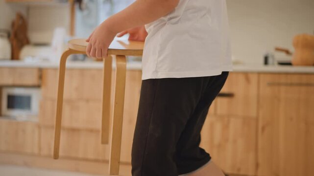 Hands carry wooden stool across kitchen, person walks with stool then bends to place seat beside warm wooden cabinets and countertop, sunlight fills room, casual clothing and socks, everyday morning