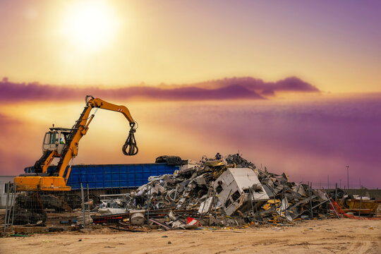 Grapple excavator moving metallic waste at an industrial recycling facility under a vibrant sky
