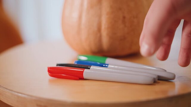 Side pumpkin on wooden stool hand reaches markers then removes them, closeup sequence showing finger grip and marker placement, warm natural tones, playful craft preparation for seasonal decoration,