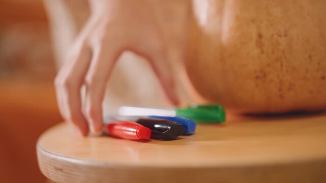 Close up pumpkin on wooden stool, hand arranging colorful markers in warm light, cozy home craft scene, tactile focus on marker stack near pumpkin, shallow depth, blurred kitchen background, intimate