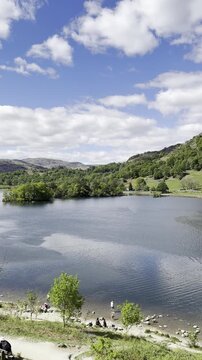Scenic view footage of Rydal Water with walking path, green hills and trees under a blue sky with scattered clouds, Lake District, England, UK.
