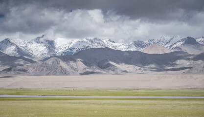 Panorama of a landscape in the Tien Shan mountains in the Pamirs, high-altitude pastures and fields with green grass against a backdrop of rocky mountains, ridges, and snow-capped peaks © Denis