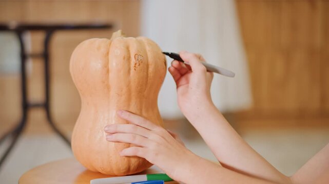 Closeup hands drawing pumpkin with marker, intimate kitchen scene with warm wood backdrop, careful outlining of template on large gourd, preparing for seasonal decoration or carving, soft daylight,
