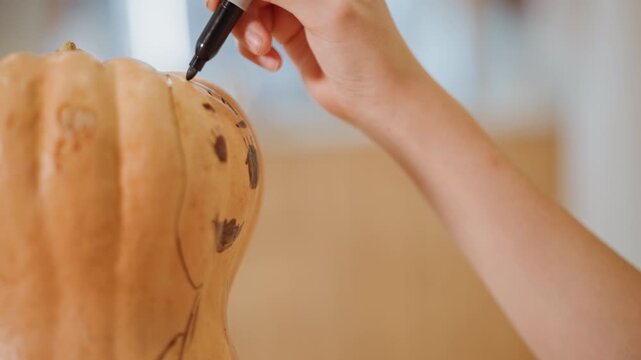 Closeup hands sketching pumpkin with marker outlining whimsical face on ribbed gourd surface, soft warm light, studio kitchen backdrop, careful artisan gesture, seasonal Halloween craft tutorial mood
